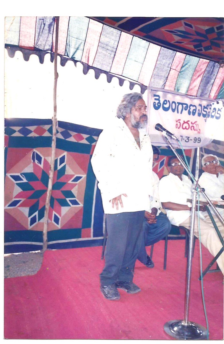 A 1999 Telangana meeting under a striped tent: Gaddar performs while Prof. Jayashankar sits on the dais
