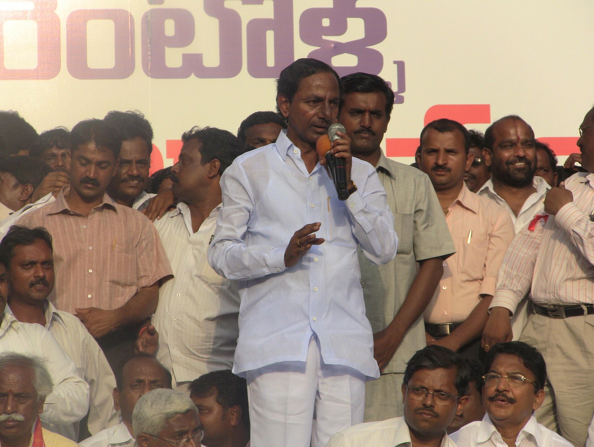 KCR speaking into a microphone at a public gathering during the Telangana agitation, surrounded by supporters