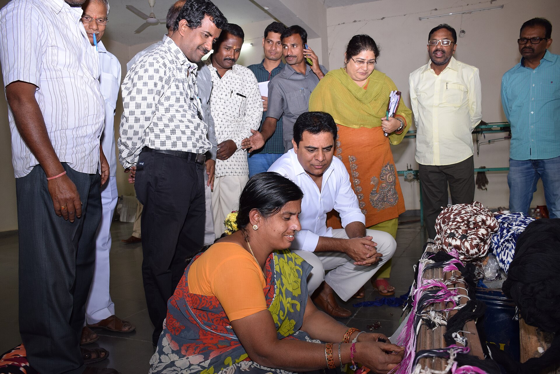 A weaver working at her loom while Minister K.T. Rama Rao crouches beside her, observing the handloom work