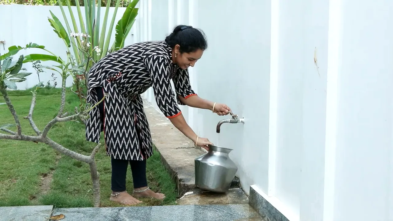 A woman filling a steel pot from a household tap beside a garden
